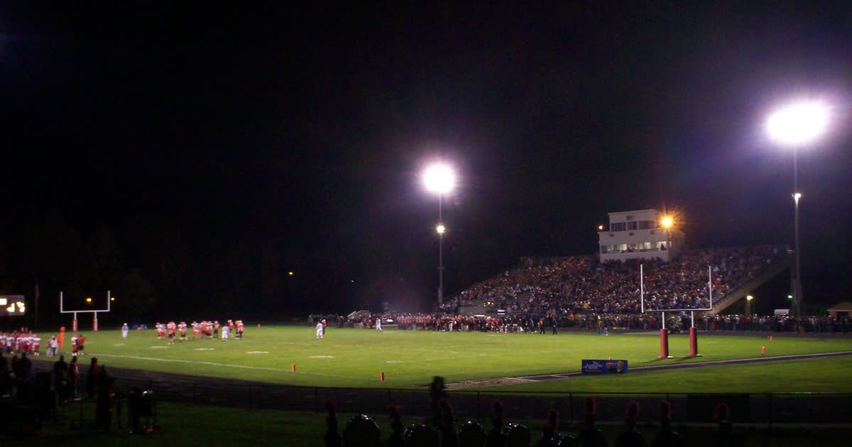 High school football stadium at night under bright lights with a full grandstand and teams on the field during a Friday night game in Ohio