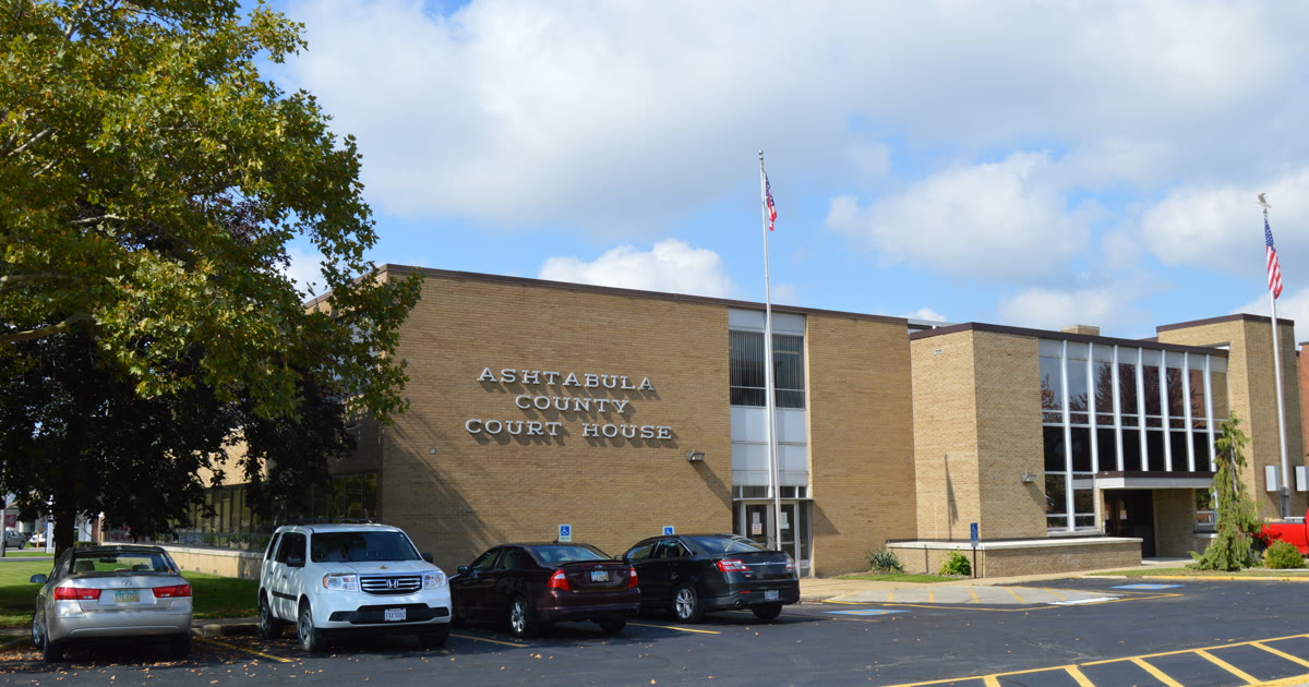Ashtabula County Courthouse in Jefferson, Ohio, its name in raised letters on the tan brick facade, with American flags and cars in the parking lot