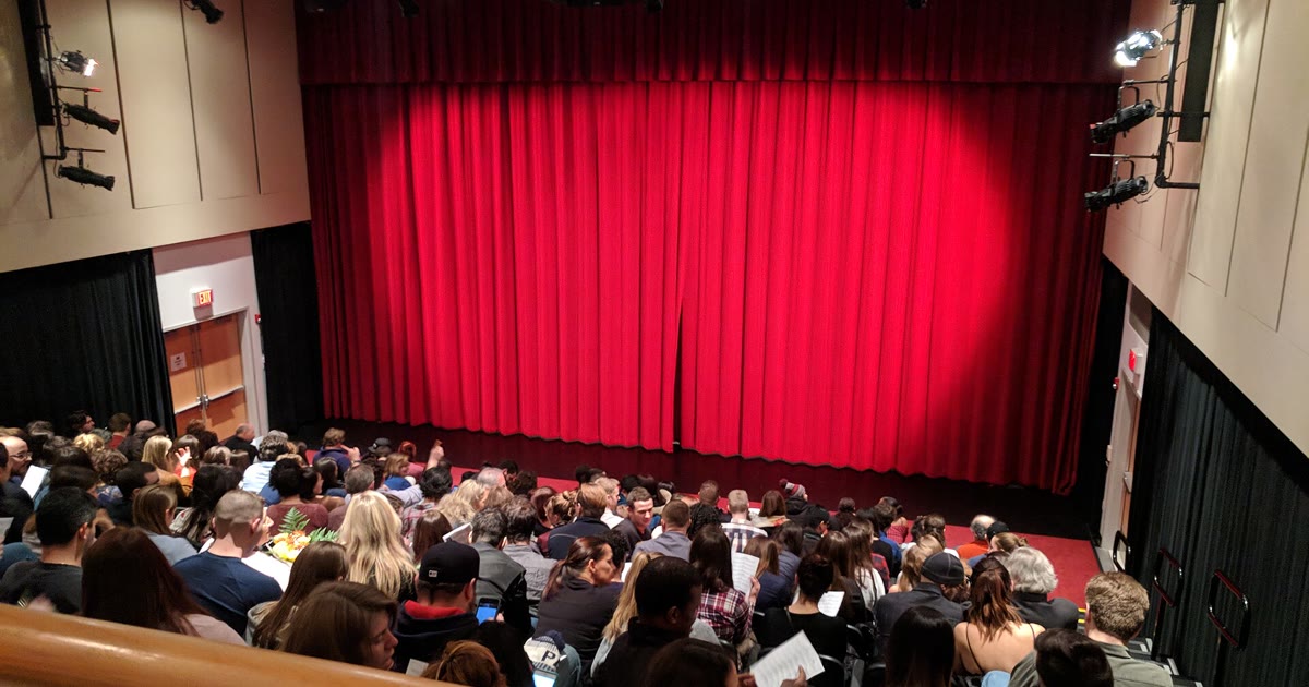An audience faces a red stage curtain in a theatre