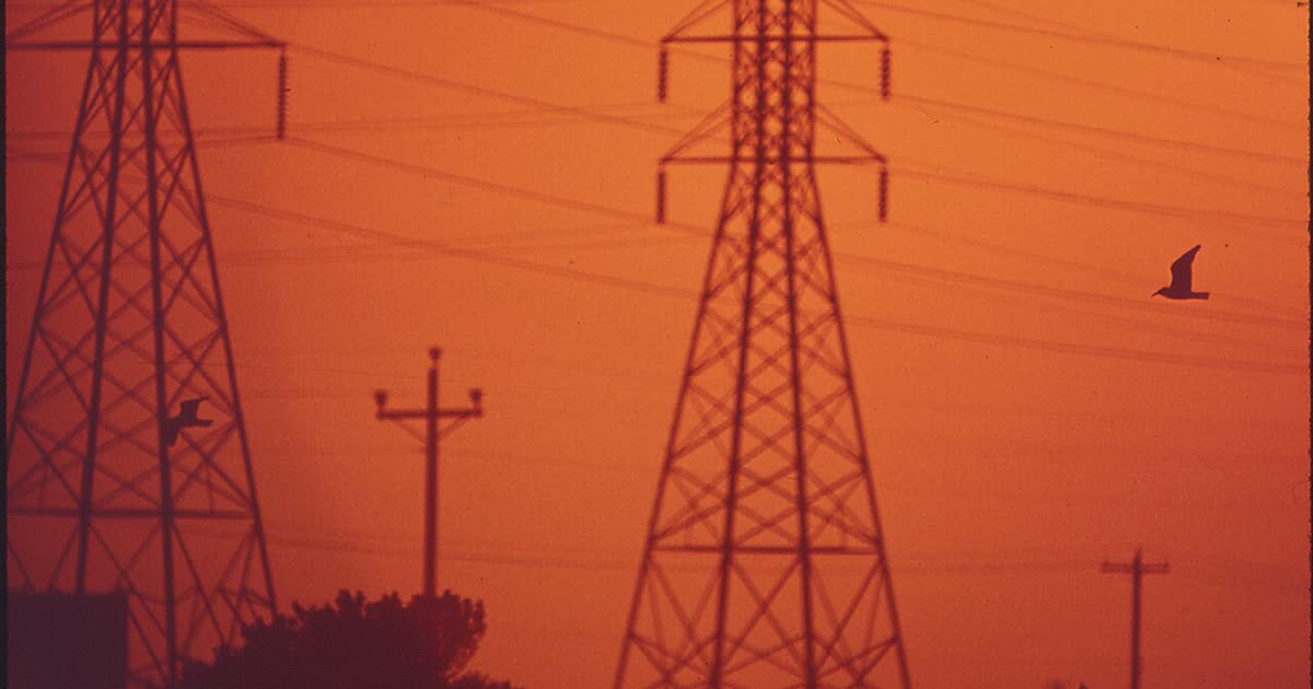 Electric transmission towers and power lines silhouetted against an orange sunset sky