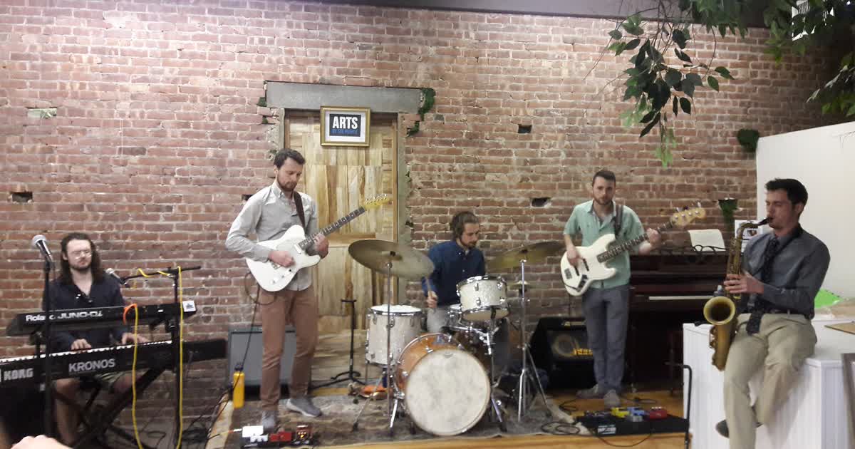 A jazz ensemble performs live on a small indoor stage with keyboard, guitars, drums and saxophone