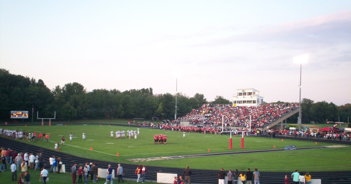 High school football teams on the field at dusk with a packed home stand under the lights